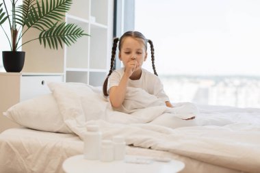 Hasta genç bayan battaniyenin altında beyaz bir yatakta oturuyor ve öksürürken ağızlarını eliyle kapatıyor. Evde otururken virüs enfeksiyonundan muzdarip beyaz saçlı küçük bir kız..