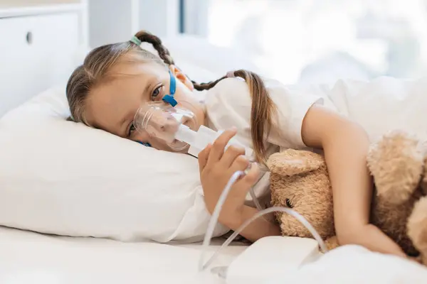 Portrait of little sad child lying in white bed and hugging bear toy while inhaling medication through nebulizer face mask. Upset caucasian girl resting and treating in bright bedroom.