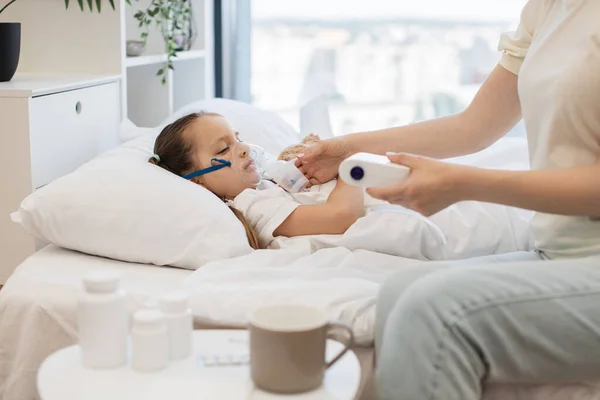 Portrait of caucasian little daughter lying in bed with toy bear and inhaling mist from nebulizer with help of caring mother. Caucasian woman supporting her sick daughter with maternal care at home.