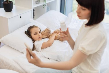 İlgilenen anne, doktorla modern akıllı telefondan iletişim kuruyor ve hasta kızının aldığı hapları gösteriyor. Tatlı kız yatakta uzanıyor ve yardım beklerken oyuncak ayıya sarılıyor..