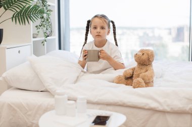 Mutsuz beyaz kız sıcak bir içecekle rahat bir yatakta dinleniyor çünkü içeride boğaz ağrısı var. Evde lezzetli çayla tatsız haplar yutmaya çalışan sevimli bir çocuk..