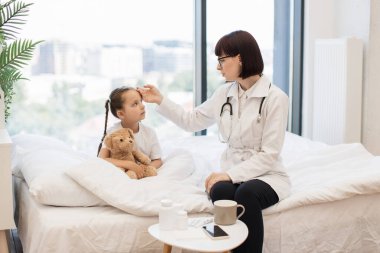 Değer veren kadın terapist vücut ısısını belirlemek için küçük bir kızın alnına elini koyuyor. Sevimli bebek en sevdiği oyuncak ayıyı kucaklıyor ve kendini yetenekli bir çocuk doktorunun ellerinde hissediyor..