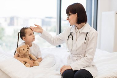 Değer veren kadın terapist vücut ısısını belirlemek için küçük bir kızın alnına elini koyuyor. Sevimli bebek en sevdiği oyuncak ayıyı kucaklıyor ve kendini yetenekli bir çocuk doktorunun ellerinde hissediyor..