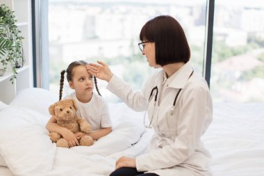 Küçük kız hasta ve yorgun hissediyor bu yüzden dost canlısı esmer hemşireden vücut ısısını kontrol etmesini istiyor. Doktorun sağlık kontrolü sırasında en sevdiği oyuncak ayıyı kucaklayan beyaz bir çocuk..