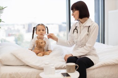 Ağızda termometreyle ısıyı ölçen, gülümseyen küçük bir kız onu tutmaya yardım ediyor. Ateşli beyaz çocuk rahat yatağında peluş ayıcığın kucağında oturuyor..