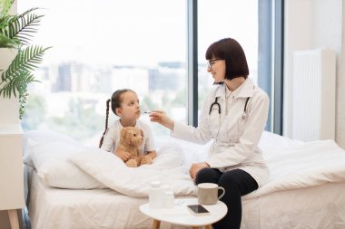 Hasta küçük kız rahat bir yatakta dinlenirken çalışkan doktor detaylı sağlık değerlendirmesi yapıyor. Kafkas çocuk doktoru çocuk sıcaklığını kontrol etmek için elektronik termometre kullanıyor..