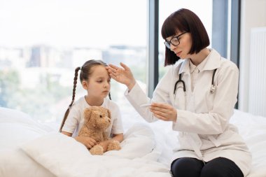 Genç hasta, beyaz yatakta, doktorun gözetiminde. Kapsamlı bir muayene yapıyor ve ateşi kontrol ediyor. Sevimli çocuk oyuncağını kucaklıyor ve ilaç almaya hazırlanıyor..