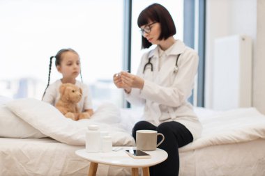 Çekici esmer çocuk doktoru elinde elektronik termometreyle hasta kızın sıcaklığını ölçüyor. Gülümseyen kadın küçük bir çocuğa ilaç yazmayı düşünüyor..