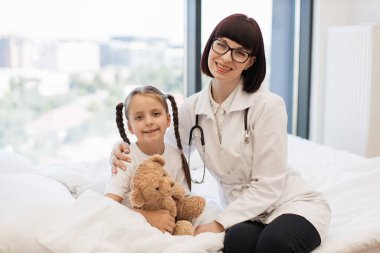 Siyah steteskoplu neşeli bir kadının portresi laboratuvar önlüğü giymiş elinde oyuncak ayıyla küçük sevimli bir kıza sarılıyor. Profesyonel tedavinin ardından başarılı bir iyileşme.