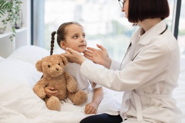 Kadın hastane çalışanı beyaz yatakta oturuyor ve elleri yatak odasında olan hasta küçük bir kızın boynuna dokunuyor. Üniformalı aile doktoru evdeki hastanın lenf düğümlerini inceliyor..