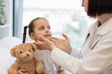 Kadın hastane çalışanı beyaz yatakta oturuyor ve elleri yatak odasında olan hasta küçük bir kızın boynuna dokunuyor. Üniformalı aile doktoru evdeki hastanın lenf düğümlerini inceliyor..