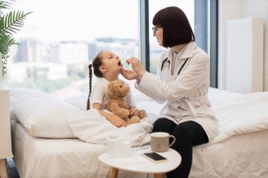 Beyaz yatakta oturan genç bir kız, sıcak yatak odasında boğazını muayene eden şefkatli bir kadın doktor. Kafkasyalı yorgun çocuk acı çekiyor ve doktor gözetiminde tedaviye ihtiyacı var..