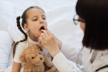 Tecrübeli bir kadın doktor, sağlıksız bir kızın boğazını içeride tıbbi aletlerle inceliyor. İş ziyareti sırasında küçük bir hastanın boğazını kontrol eden laboratuvar önlüklü beyaz kadın..