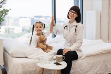 Güzel beyaz çocuk doktoru ve sevimli küçük kız yatakta otururken beşlik çakıyor. Doktor ve hastalardan oluşan dost canlısı bir ekip evdeki tedavi sonuçlarından memnun..