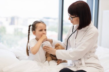 Steteskoplu küçük sevimli bir çocuğun nefes alan organlarını dinleyip öksürmesini isteyen nitelikli beyaz bir hemşire. Dost canlısı doktor küçük kızı muayene ediyor. Rahat yatak odasında tedavi için..