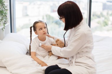 Modern yatak odasının panoramik penceresinden anaokulu çocuğunun solunum sistemini kontrol etmek için steteskop kullanan beyaz bir hemşire. Tedavi, pediatri ve sağlık kavramı.
