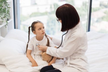 Hasta bir kızı yatağa yatırıp steteskopla muayene eden yetişkin bir kadın. Küçük çocuk kötü bir şekilde dinleniyor ve elinde oyuncakla evde doktorun muayenesini bekliyor..