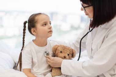 Hasta bir kızı yatağa yatırıp steteskopla muayene eden yetişkin bir kadın. Küçük çocuk kötü bir şekilde dinleniyor ve elinde oyuncakla evde doktorun muayenesini bekliyor..