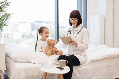 Geniş yatak odasındaki hasta çocuk için modern tablete ilaç yazan becerikli bir kadın pediyatrist. Gülümseyen kız oyuncak ayıyı tutuyor ve evde yoğun tedaviye hazırlanıyor..