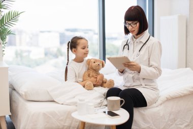 Dijital tabletle yazı yazan deneyimli bir kadın pediatrist ev ziyareti sırasında küçük hasta hasta hakkında detaylı bilgi verdi. Kafkasyalı yorgun kız peluş ayısıyla sarılıp yatıyor..