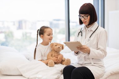 Dijital tabletle yazı yazan deneyimli bir kadın pediatrist ev ziyareti sırasında küçük hasta hasta hakkında detaylı bilgi verdi. Kafkasyalı yorgun kız peluş ayısıyla sarılıp yatıyor..