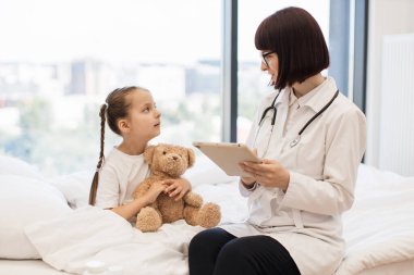 Sayısal tabletli çekici hasta kızın yanında rahat bir yatakta oturan nitelikli bir doktor. Dost canlısı beyaz kadın kişisel bilgilerini çevrimiçi tıbbi karta aktarıyor..