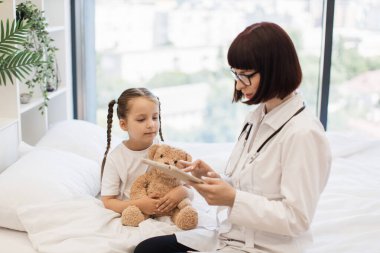 Sayısal tabletli çekici hasta kızın yanında rahat bir yatakta oturan nitelikli bir doktor. Dost canlısı beyaz kadın kişisel bilgilerini çevrimiçi tıbbi karta aktarıyor..