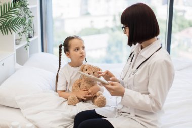 Sayısal tabletli çekici hasta kızın yanında rahat bir yatakta oturan nitelikli bir doktor. Dost canlısı beyaz kadın kişisel bilgilerini çevrimiçi tıbbi karta aktarıyor..