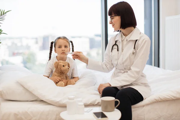 Upset small girl measuring temperature with thermometer in mouth while smiling doctor helping to holding it. Adorable caucasian kid with fever sitting on comfy bed in embrace of plush teddy bear.