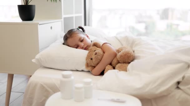 疲れた白人少女は柔らかいベッドに横たわり テディベアを抱擁し 病気を感じています 近くに立っている丸薬と温度計が付いているコーヒー テーブル 医療と病気のコンセプト — ストック動画