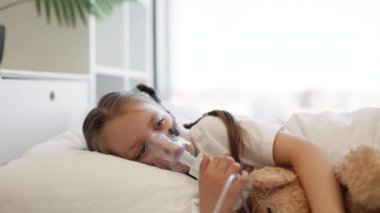 Küçük üzgün bir çocuğun portresi beyaz yatakta yatarken ve ayı oyuncağına sarılırken nebulizör maskesiyle ilaç soluyor. Kafkasyalı üzgün kız dinleniyor ve aydınlık yatak odasında tedavi oluyor..