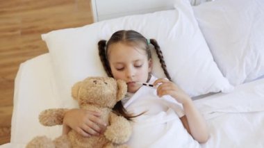 Hasta küçük kızın, yatakta ağzında dijital termometreyle sevgili oyuncak ayısına sarılışının en üst görüntüsü. Tıbbi hastalık ve ateş kavramı.