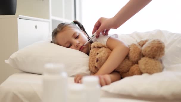 病気の女の子の口に電子温度計を置く女性の手の近く 疲れた慎重な子供はテディベアのおもちゃでベッドに横たわり 母親は彼女の温度を測定しています — ストック動画