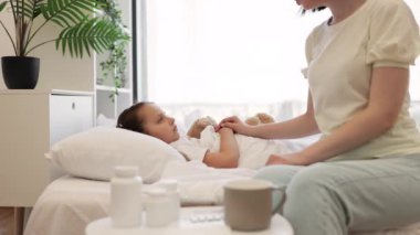 Beyaz yatakta hasta kızının yanında oturan, oyuncak ayıyı kucaklayan ve sıcaklığı elle ölçen, şefkatli beyaz bir annenin görüntüsü. Aile ve sağlık durumu kavramı.