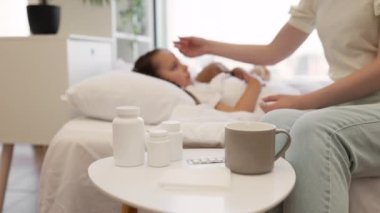 Beyaz yatağın yanında duran sıcak içecek, kağıt mendil ve ilaç dolu komodinlere odaklan. Kızların ateşini dijital termometreyle ölçen ilgili annenin bulanık arkaplanı..