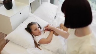 Beyaz yatakta oturan, dijital termometreye bakan ve çocuk alnında elini tutan beyaz saçlı kadın. Hasta kızının evde sıcaklığını kontrol eden ilgili bir anne..