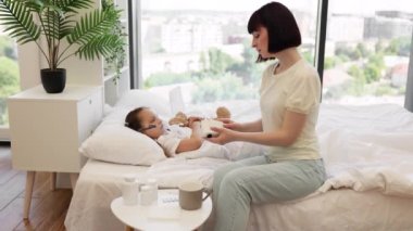 Sıradan giyinen beyaz kadın, hasta kızının evde nebulizer solunumu ile hastalığın üstesinden gelmesine yardım ediyor. Sevecen bir anne hasta çocuğunun yanında oturuyor ve aile içi bakım sağlıyor..