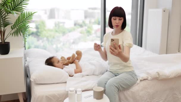 現代のスマートフォンで小児科医とビデオ通話をする母親は 自宅で不健康な娘を治療しています 疲れた少女はテディベアのおもちゃを抱擁し 毛布の下でベッドに横たわる — ストック動画