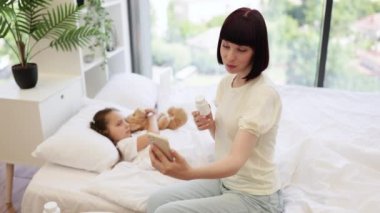 Çekici beyaz kadın, küçük kızına davranılmasından bahsederken kişisel akıllı telefonuyla görüntülü konuşma yapıyor. Güzel çocuk rahat bir yatakta uzanıyor ve dikkatlice annesini dinliyor..