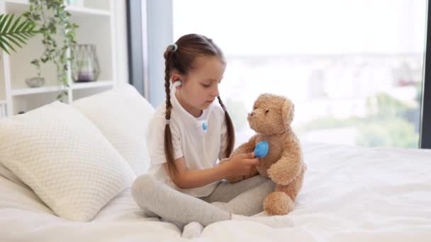 小女孩用玩具听诊器在舒适的床上 在城市全景窗的背景下 听着玩具熊的心跳 游戏和治疗的概念 — 图库视频影像