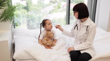 Nazik beyaz esmer doktor, küçük bir çocuğun sıcak alnına yaslanarak termometre ölçümlerini kontrol ediyor. Yorgun kız iyileşmek için en sevdiği oyuncak ayıyı kucaklıyor..
