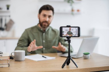 Modern akıllı telefon tripoda tamir edildi. Günlük giyim giyen yakışıklı beyaz adam ofis bilgisayarında çalışıyor. Arka plan bulanık. Teknoloji, insanlar ve blog kavramı.