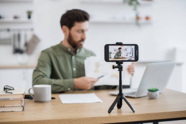 Masasında dizüstü bilgisayarla oturan ve akıllı telefon kamerasında mali raporunu gösteren beyaz adamın bulanık arkaplanı. Uzman finansör grafik grafikleri okumak ve yorumlamak hakkında video dersi veriyor.