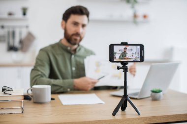 Masasında dizüstü bilgisayarla oturan ve akıllı telefon kamerasında mali raporunu gösteren beyaz adamın bulanık arkaplanı. Uzman finansör grafik grafikleri okumak ve yorumlamak hakkında video dersi veriyor.