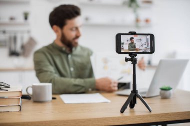 Masasında dizüstü bilgisayarla oturan ve akıllı telefon kamerasında mali raporunu gösteren beyaz adamın bulanık arkaplanı. Uzman finansör grafik grafikleri okumak ve yorumlamak hakkında video dersi veriyor.