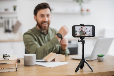 Canlı yayın sırasında elinde elektronik siyah saati olan aktif bir adamın bulanık arka planı. İlgilenen erkek nüfuzlu kişi güncel aygıtı takıyor ve sayfasında yorum istiyor.