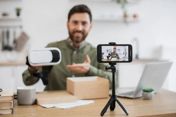 Happy caucasian man showing virtual reality glasses while filming himself of modern smartphone standing on tripod. Handsome technology blogger working remotely in personal cabinet at home.