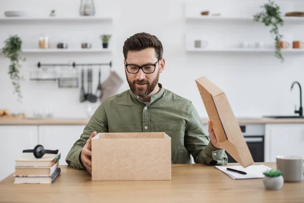 First person view of male technology blogger in eyeglasses sitting at desk and opening cardboard box. Positive bearded man creating interesting new vlog while receiving new parcel at home.