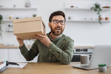 Evdeki ofiste elinde karton kutuyla oturan sıradan giysili ve gözlüklü heyecanlı bir beyaz adam. Sakallı erkek blogcu tatil indirimi sırasında online mağazadan sipariş aldığı için mutlu..