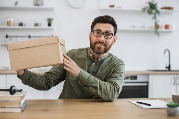 stock image Excited caucasian man in casual wear and glasses sitting at home office with cardboard box in hands. Bearded male blogger happy to receive order from online store during holiday discount.